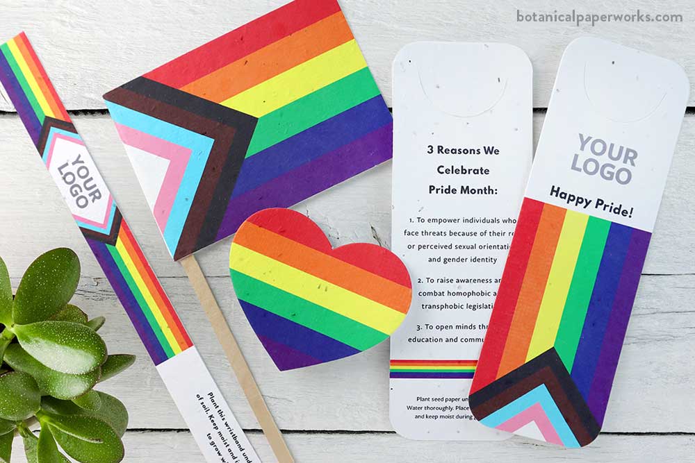 A Seed Paper Pride Flag, Plantable Pride Wristband, Plantable Pride Bookmark with Page Slot, and Plantable Pride Heart Button Bages from Botanical PaperWorks. Promotional giveaways for Pride Month and other LGBTIQ events featuring the Progress Pride Flag.