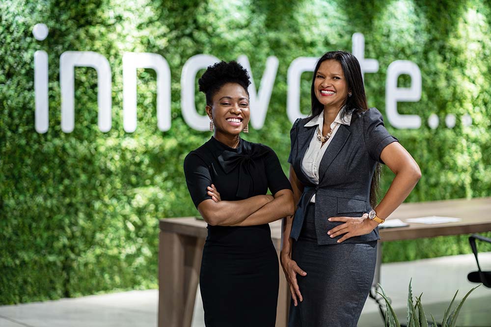 Two women standing in front of a greenery wall with the sign "innovate"