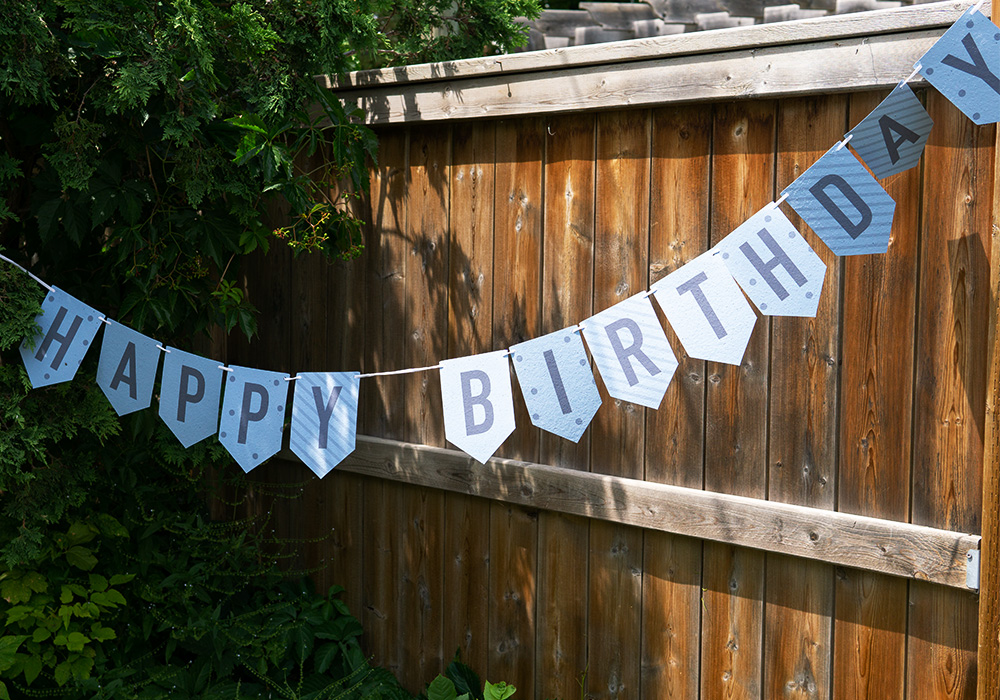Blue bunting banner that reads, happy birthday, hanging from a tree with a wooden fence in the background.