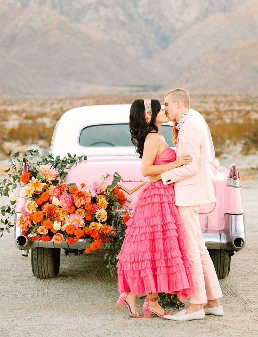 A bride and groom kissing in front of a pink classic car.