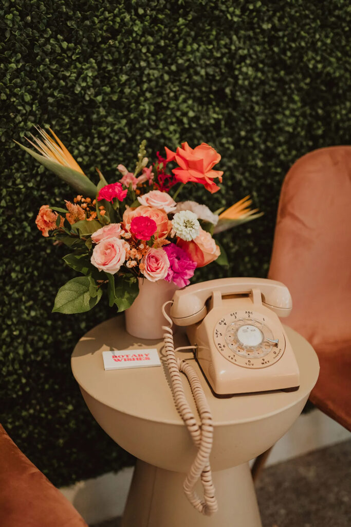 A brightly colored floral arrangment on a side table, with a old telephone next to it. 