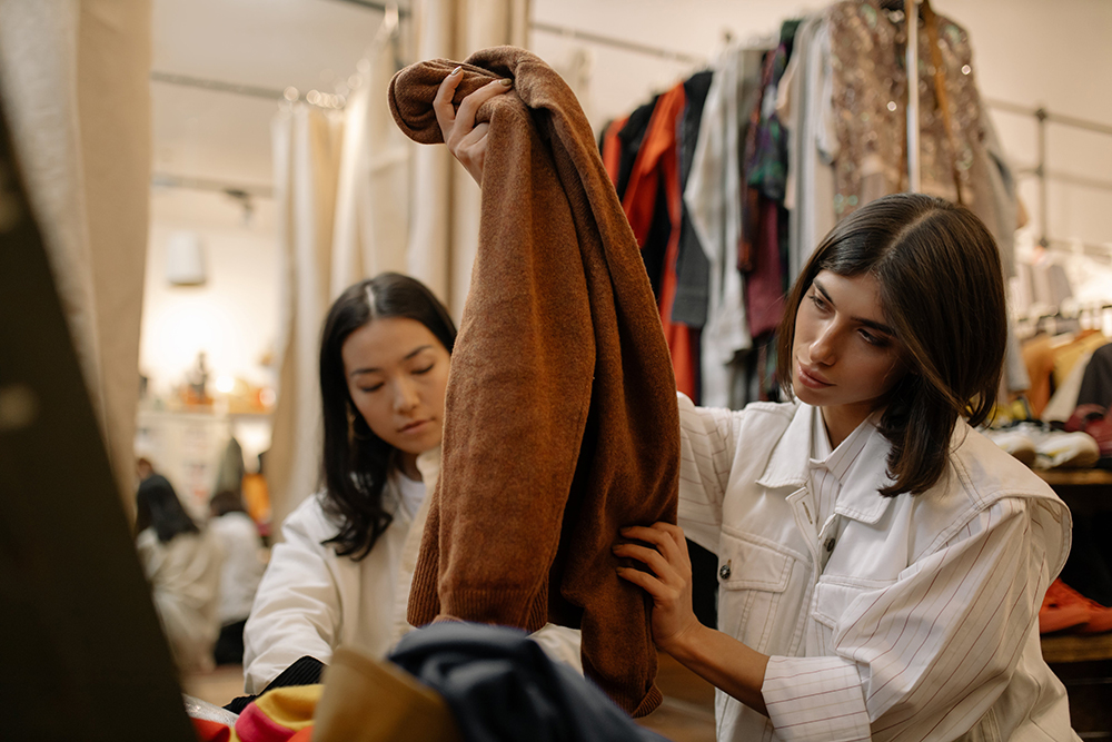 Two females looking though clothes at a thrift store. One is holding up a brown jacket. 