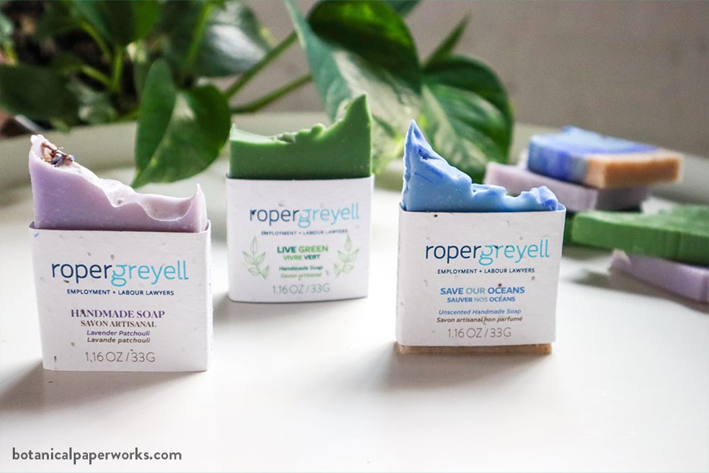 Three artisanal soaps in purple, green, and blue sit upright on a white surface, each wrapped in plantable seed paper bands branded with the Roper Greyell Employment + Labour Lawyers logo. The labels identify the scents as Lavender Patchouli, Live Green, and Save Our Oceans. Blurred soaps and green foliage appear in the background.